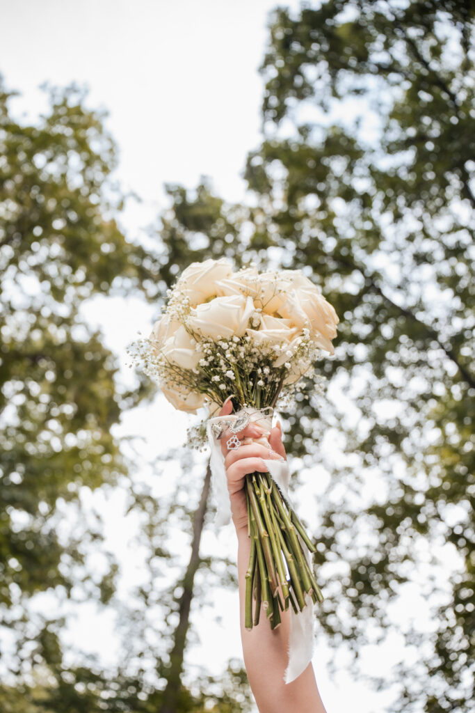 Bouquet of white roses being held in the air with a tree canopy in the background. 